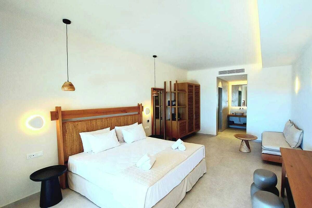 A hotel room with a large bed, a wooden headboard, a bedside table, a small round table, a sofa, and a TV mounted on the wall.