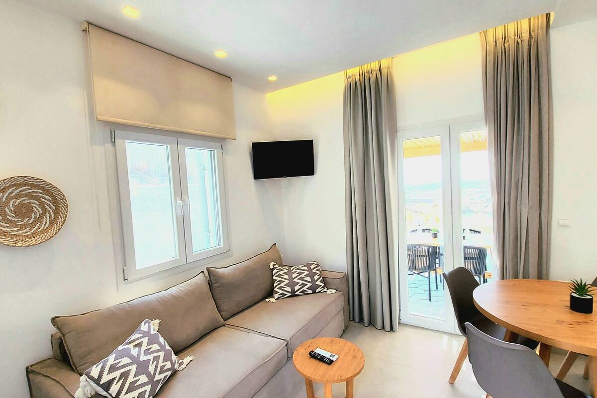 A modern living room with a sofa, a TV mounted on the wall, and a dining area with a table and chairs.