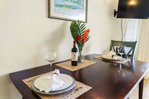 Een eettafel gedekt met borden, servetten en een fles wijn, met een lijst met een schilderij aan de muur en een televisie erboven.