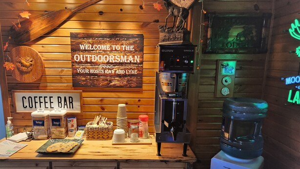 Un bar de café con un letrero que dice "WELCOME TO THE OUTDOORSMAN OUR HOSTS RAY AND LYNE" y una máquina de café.