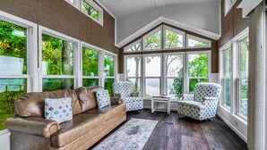 A sunroom with a leather sofa, patterned chairs, and large windows offering a view of trees. - Paradise Point- Sleeps 2 HOT TUB (De Soto)