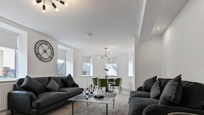 A modern living room with a black sofa, a coffee table, and a dining area with green chairs. - Mode Winter Garden Apartments (Blackpool)