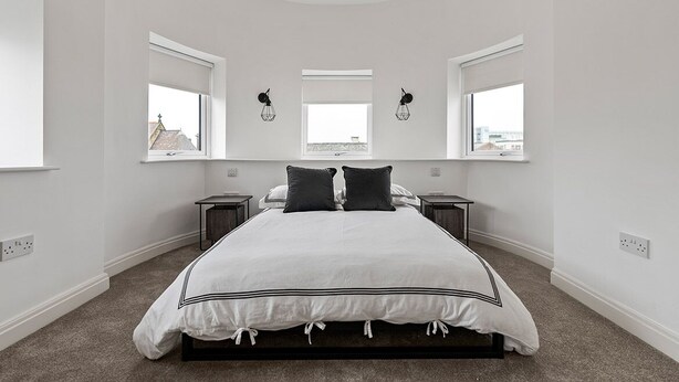 A bedroom with a large bed, two bedside tables, and three windows. - Mode Winter Garden Apartments (Blackpool)