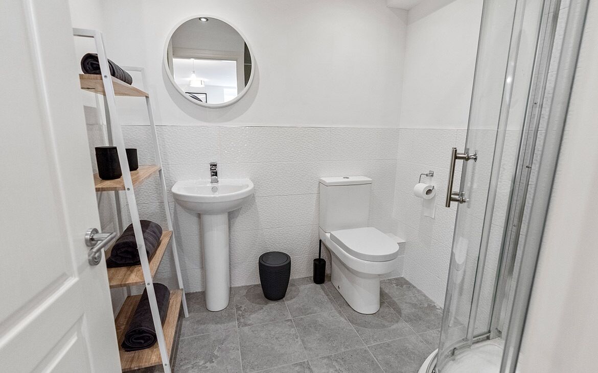 Un baño moderno con un lavamanos, inodoro y ducha blancos. Hay repisas con toallas y un bote de basura.