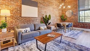 Ein Wohnzimmer mit Backsteinwand, einer Couch, einem Couchtisch, einem Esstisch mit Stühlen und einem großen Fenster.