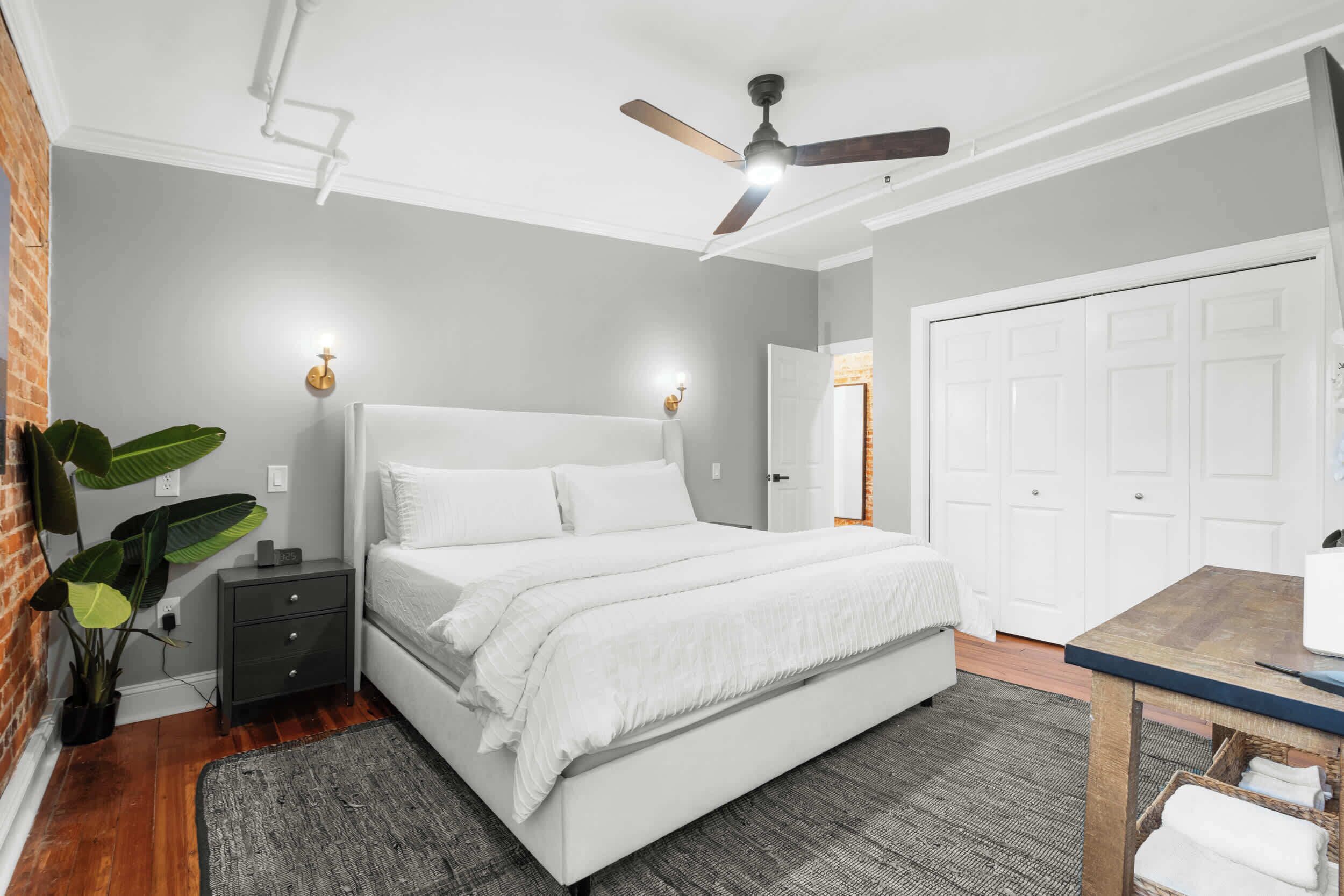 Un dormitorio con cama, mesita de noche, lámparas de noche, ventilador de techo y puerta a otra habitación.