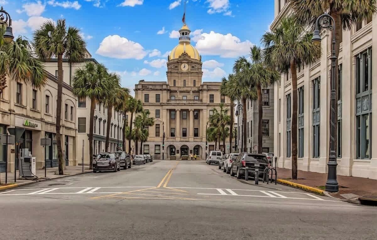 A street view of a building with a large dome and columns, surrounded by parked cars and palm trees.
