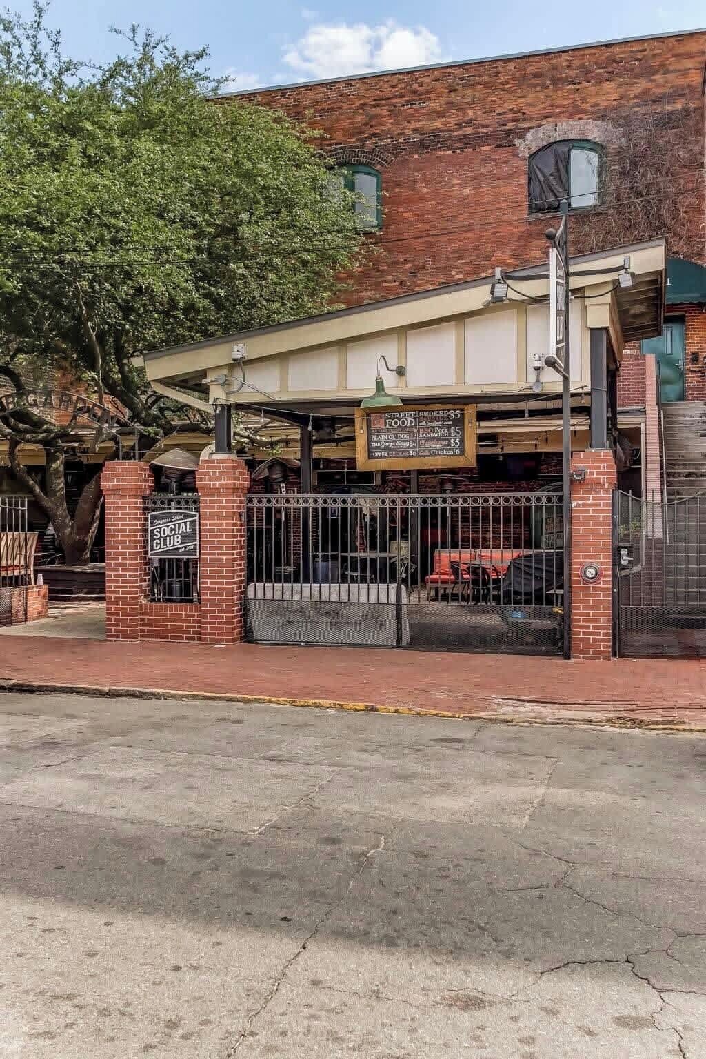 Een bakstenen gebouw met een overdekt terras, een bord met de tekst 'Street Smoked Food' en prijzen, en een bord met 'Social Club'.