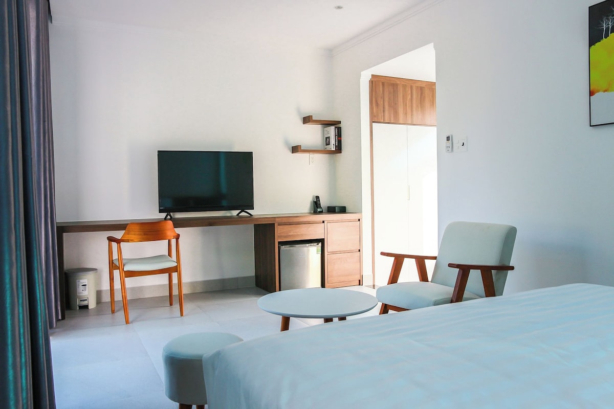 Quarto de hotel com cama, escrivaninha com televisão, cadeira, mesa pequena e lixeira.