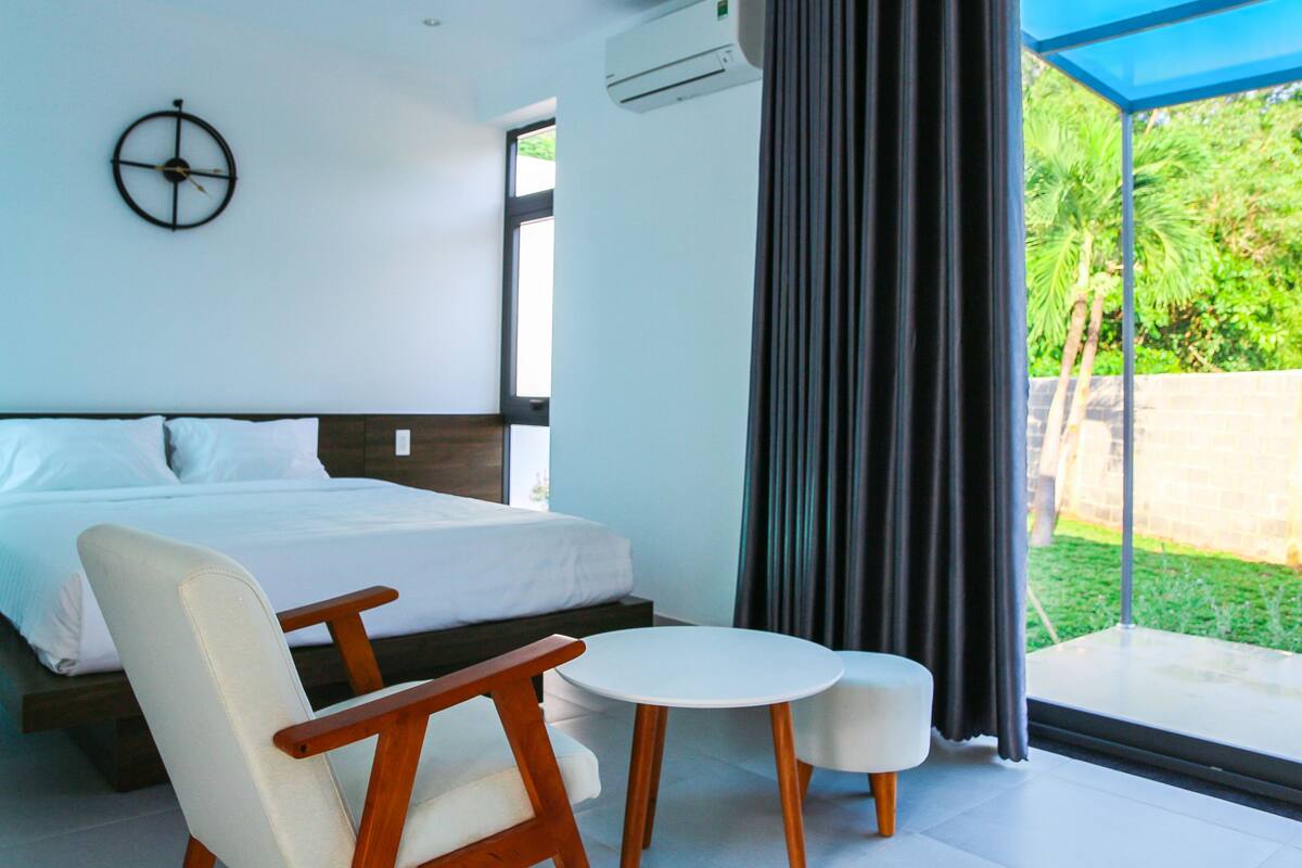 A modern hotel room with a large bed, a wooden chair, a small round table, and a view of greenery outside.