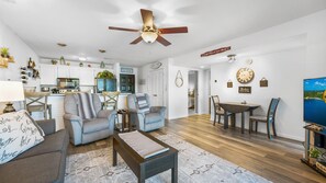 A spacious living room with a ceiling fan, a dining area with a table and chairs, a kitchen with white cabinets, and a large wall clock.