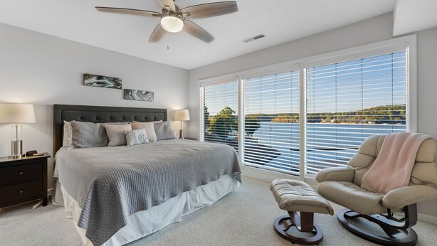 Une chambre à coucher avec un lit, des tables de chevet, une chaise et une vue sur un lac, visible à travers de grandes fenêtres.