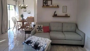 A living room with a sofa, a coffee table, a dining table with chairs, and a chandelier. There are wall shelves with decorative items. - Cozy Getaway 5 Min Walk to Parks (Royal Palm Beach)