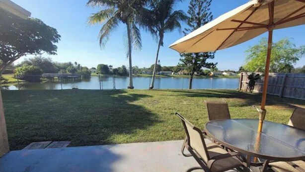 A patio with a table and chairs, a lawn, and a view of a lake with palm trees. - Cozy Getaway 5 Min Walk to Parks (Royal Palm Beach)