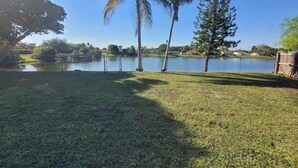 A grassy area with a body of water, palm trees, and a wooden fence. - Cozy Getaway 5 Min Walk to Parks (Royal Palm Beach)