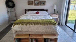 A bedroom with a bed, a wooden bench with books, a nightstand, a lamp, and a window with a view of greenery. - Cozy Getaway 5 Min Walk to Parks (Royal Palm Beach)