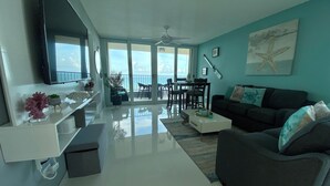 Ein modernes Wohnzimmer mit Sofa, Couchtisch und Fernseher. Ein Balkon bietet Blick auf den Ozean.