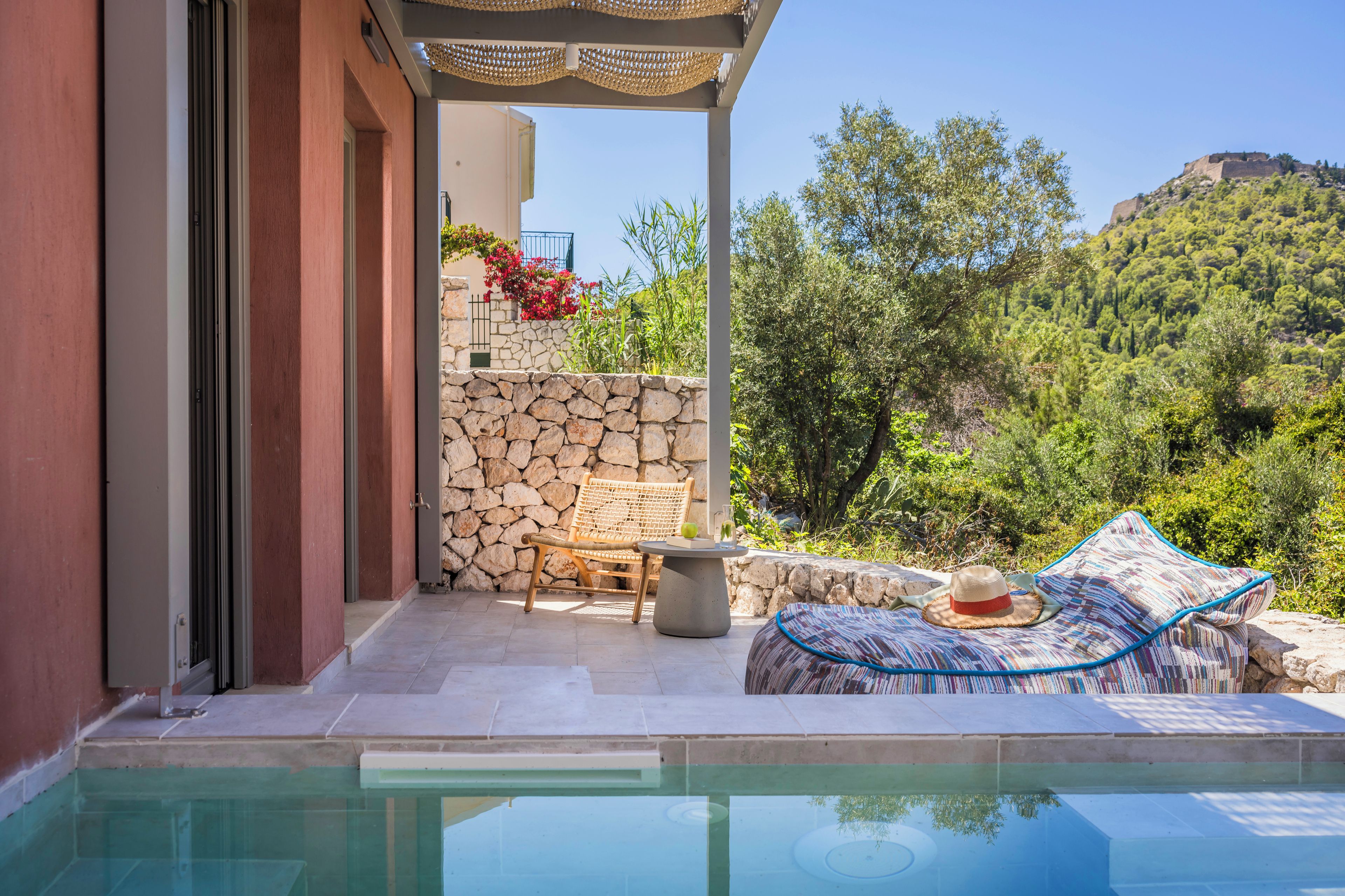A poolside area with a stone wall, a wooden bench, and a lounge chair with a hat.