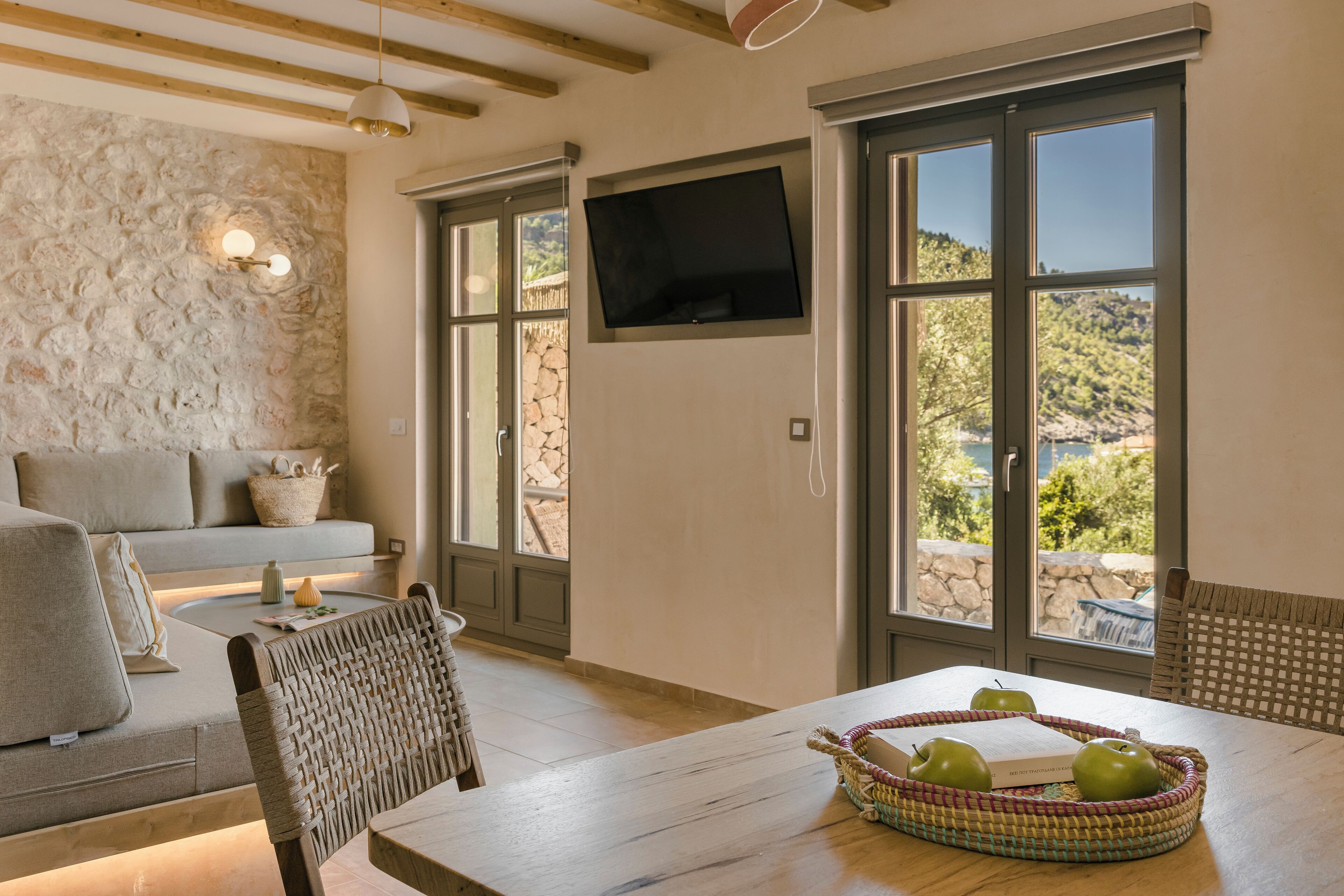 Una habitación acogedora con pared de piedra, un sofá, una mesa de comedor y una cesta de mimbre llena de manzanas, además de un televisor montado en la pared.