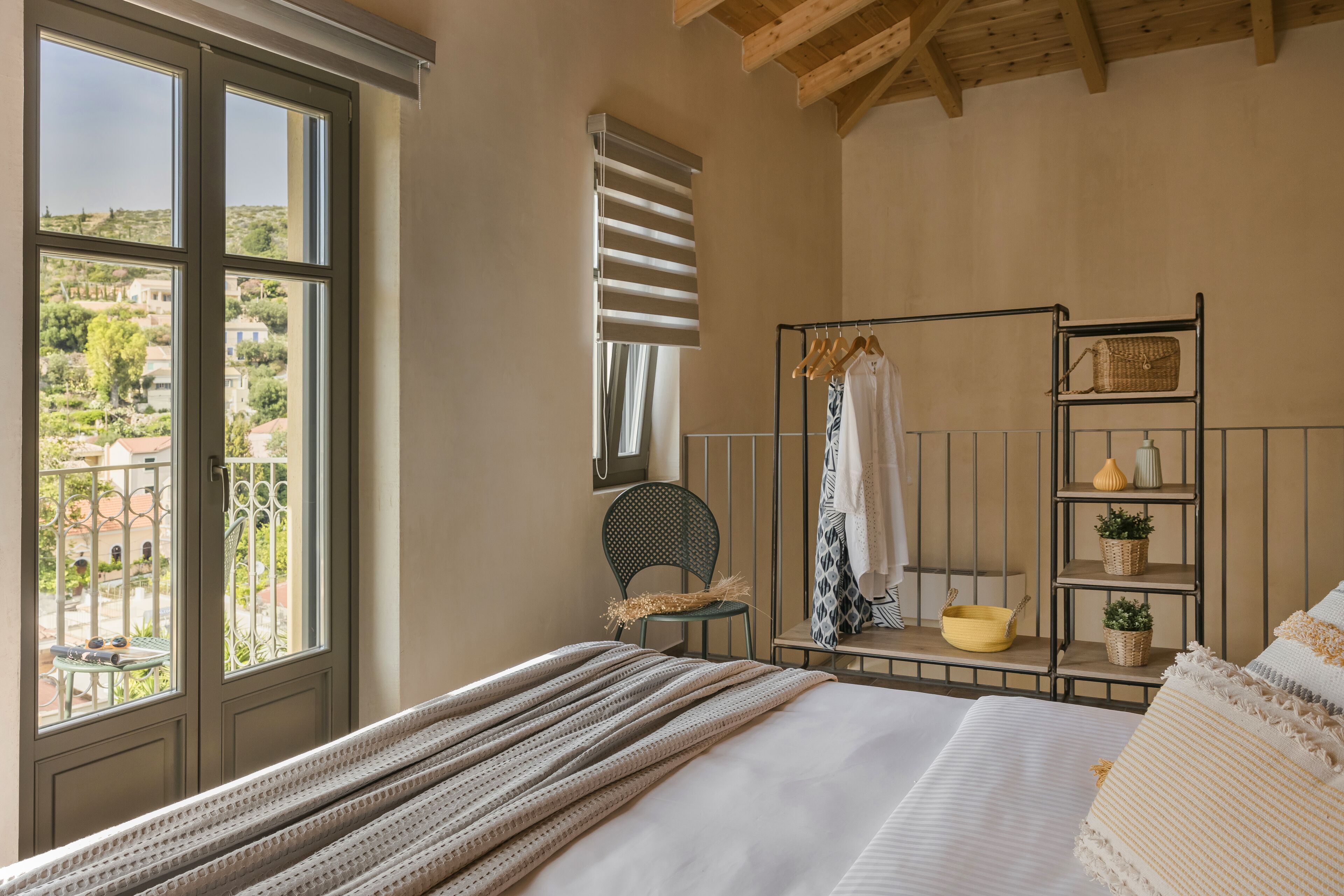 Een slaapkamer met een groot raam, een bed met een gestreept dekbed, een stoel, een metalen plankje met decoratieve objecten en uitzicht op buiten.