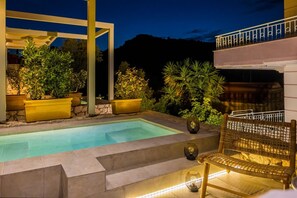 A swimming pool with steps leading into it, surrounded by potted plants and a wicker chair. - Assos Chromata Villas (Kefalonia)