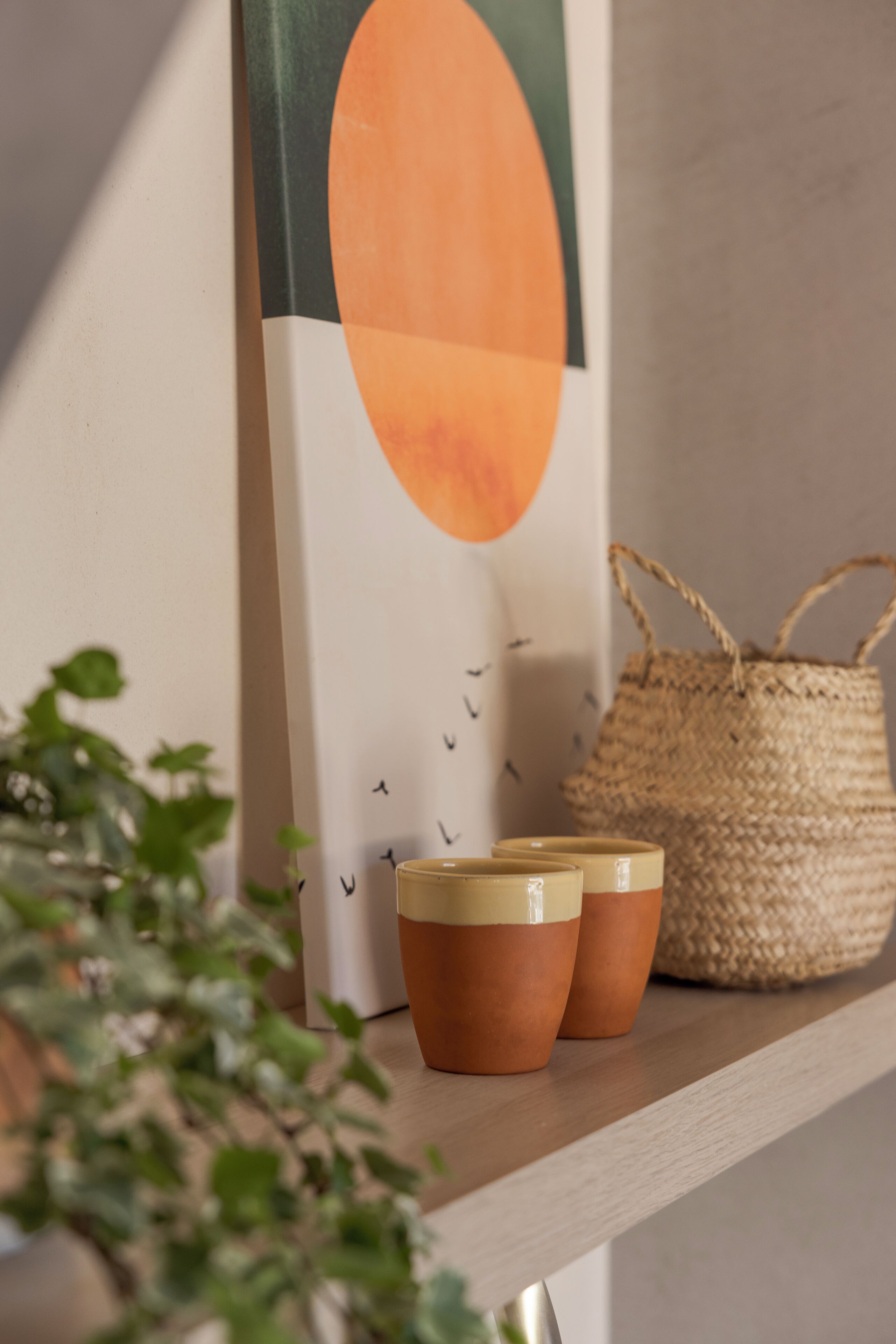 A shelf with a woven basket, two ceramic cups, and a framed artwork.