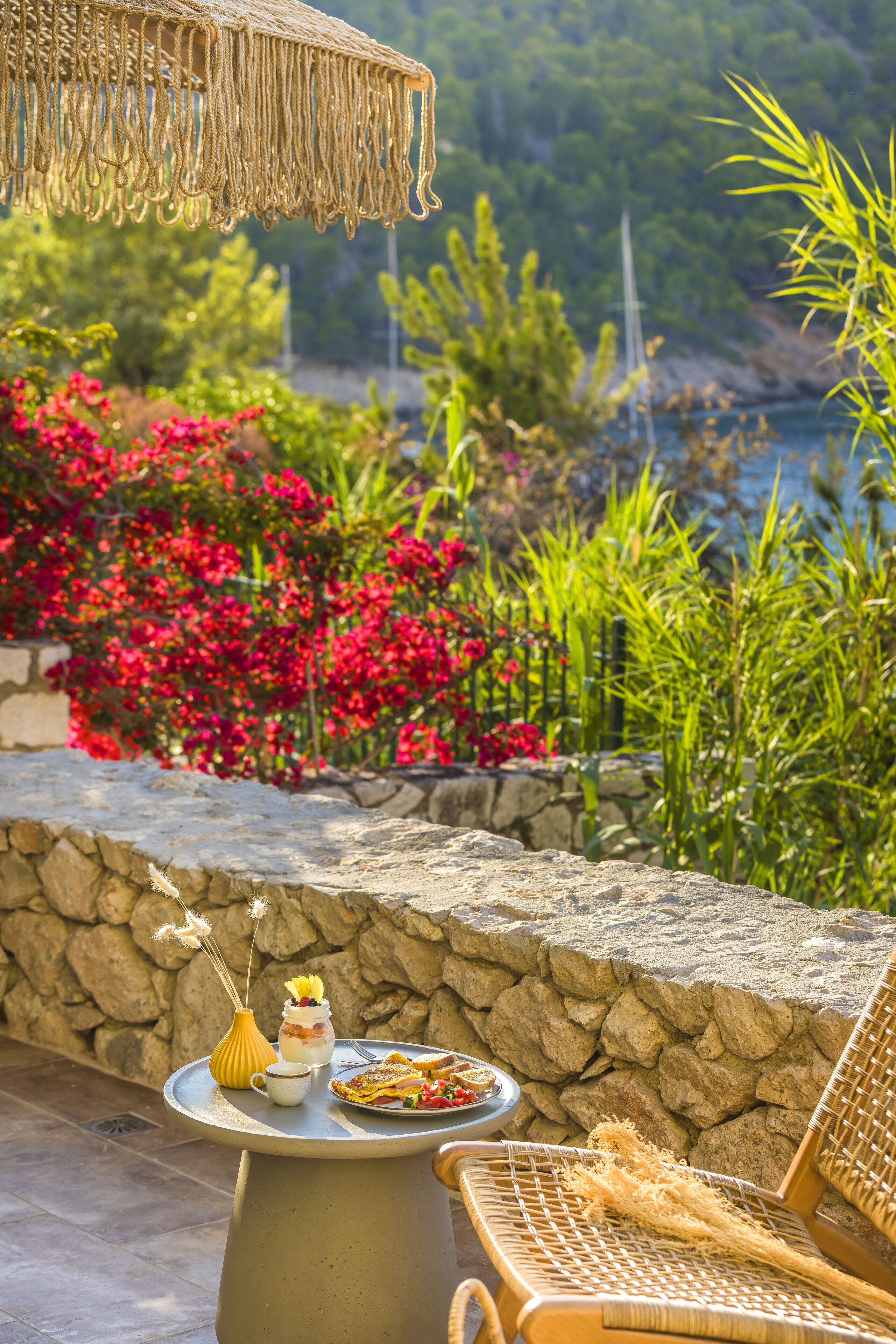 A stone wall with a table set for a meal, a wicker chair, and a view of a garden with red flowers and greenery.