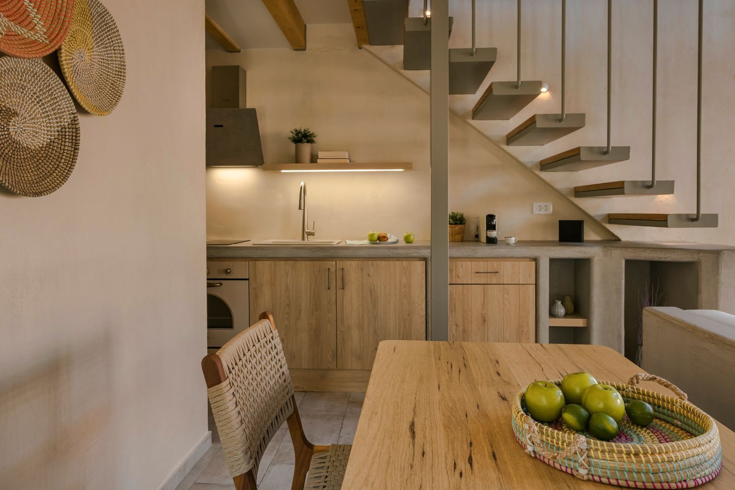 Una cocina moderna con armarios de madera, un lavamanos de acero inoxidable y una zona de comedor con mesa de madera y silla tejida.