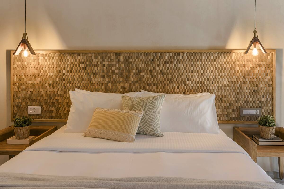 A neatly made bed with white linens and two decorative pillows, a wooden textured headboard, and two bedside tables with small potted plants.