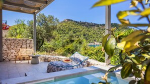 A pool area with a stone wall, a wooden chair, a cushion, and a view of a hill with a building. - Assos Chromata Villas (Kefalonia)