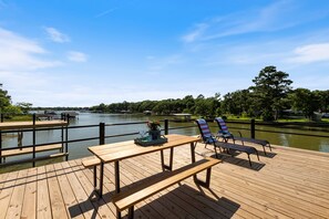 Una terraza de madera con una mesa y dos sillas, con vista a un cuerpo de agua tranquilo y una costa distante.