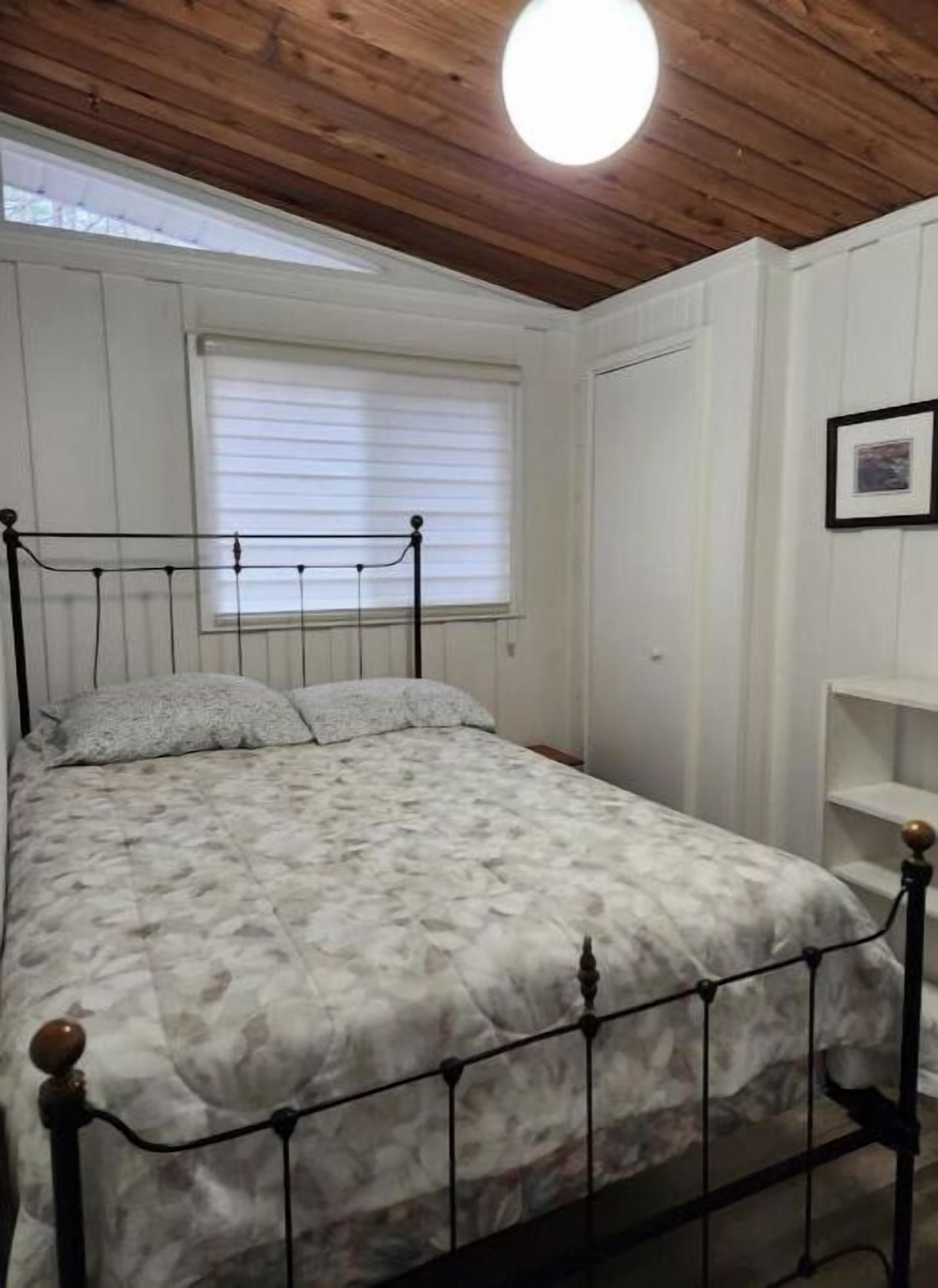 Ein Schlafzimmer mit einem Bett, einem Fenster mit Jalousien, einer Holzbalkendecke und einer weißen Wand mit einem Bilderrahmen.
