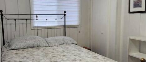 Ein Schlafzimmer mit einem Bett, einem Fenster mit Jalousien, einer Holzbalkendecke und einer weißen Wand mit einem Bilderrahmen.