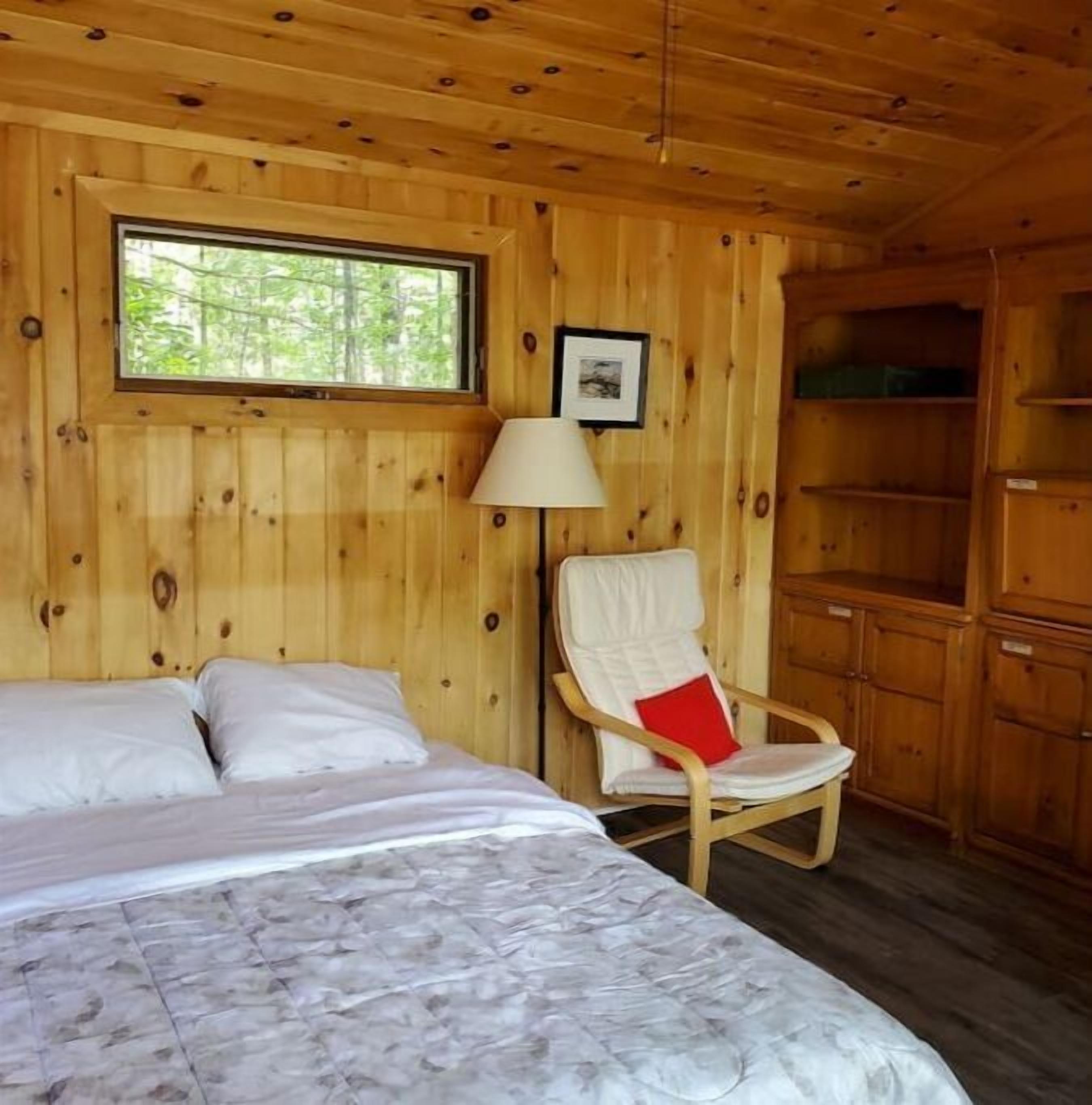 A wooden cabin room with a bed, a chair, a window, and a shelf.