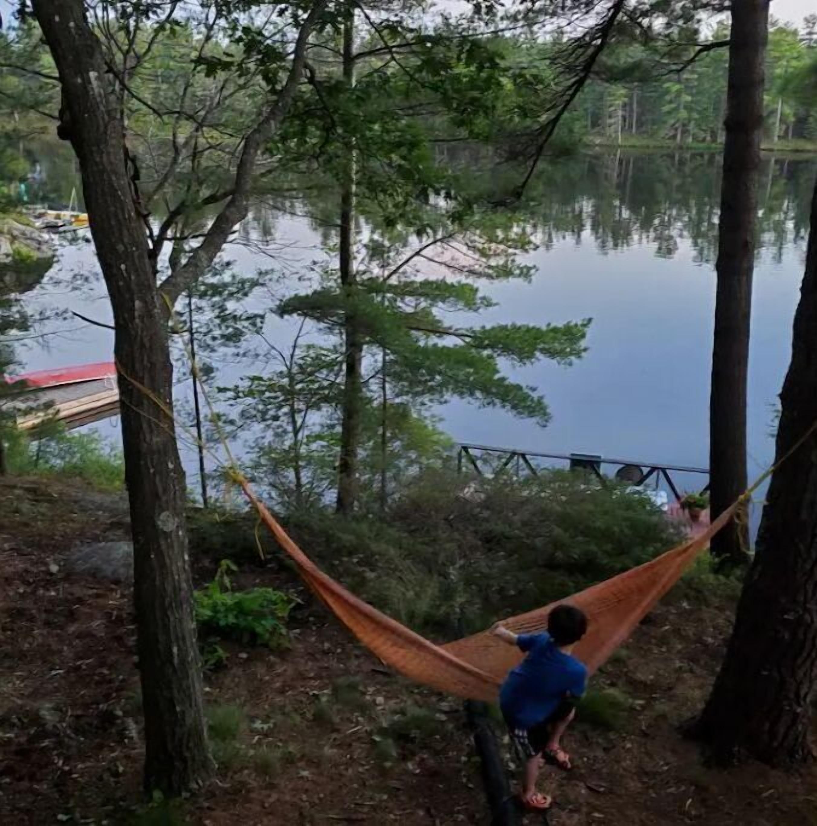 A hammock tied between trees overlooking a calm lake with a dock in the background.