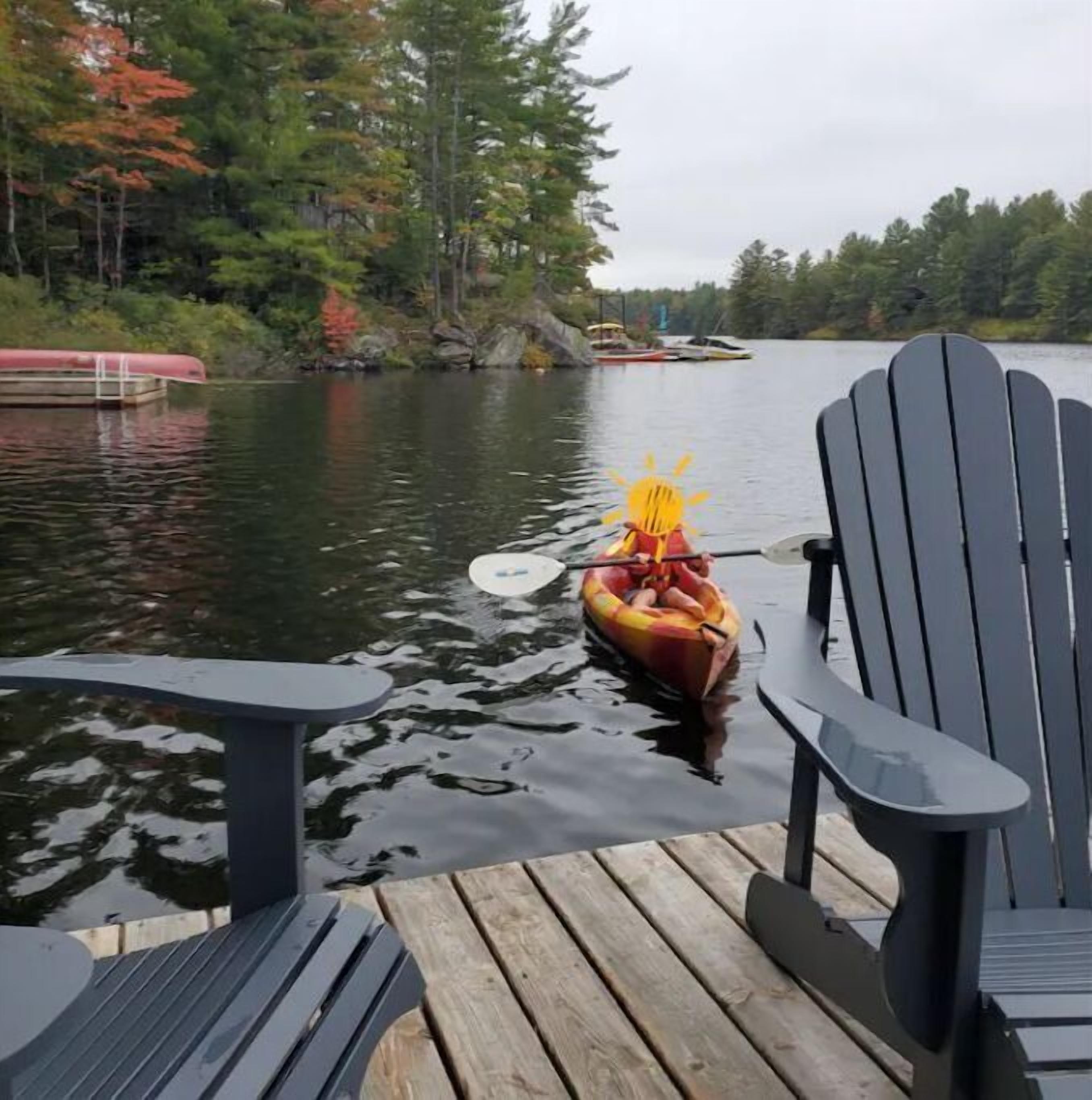 Um cais de madeira com cadeiras Adirondack, um canoa com um remo e um lago calmo rodeado por árvores.
