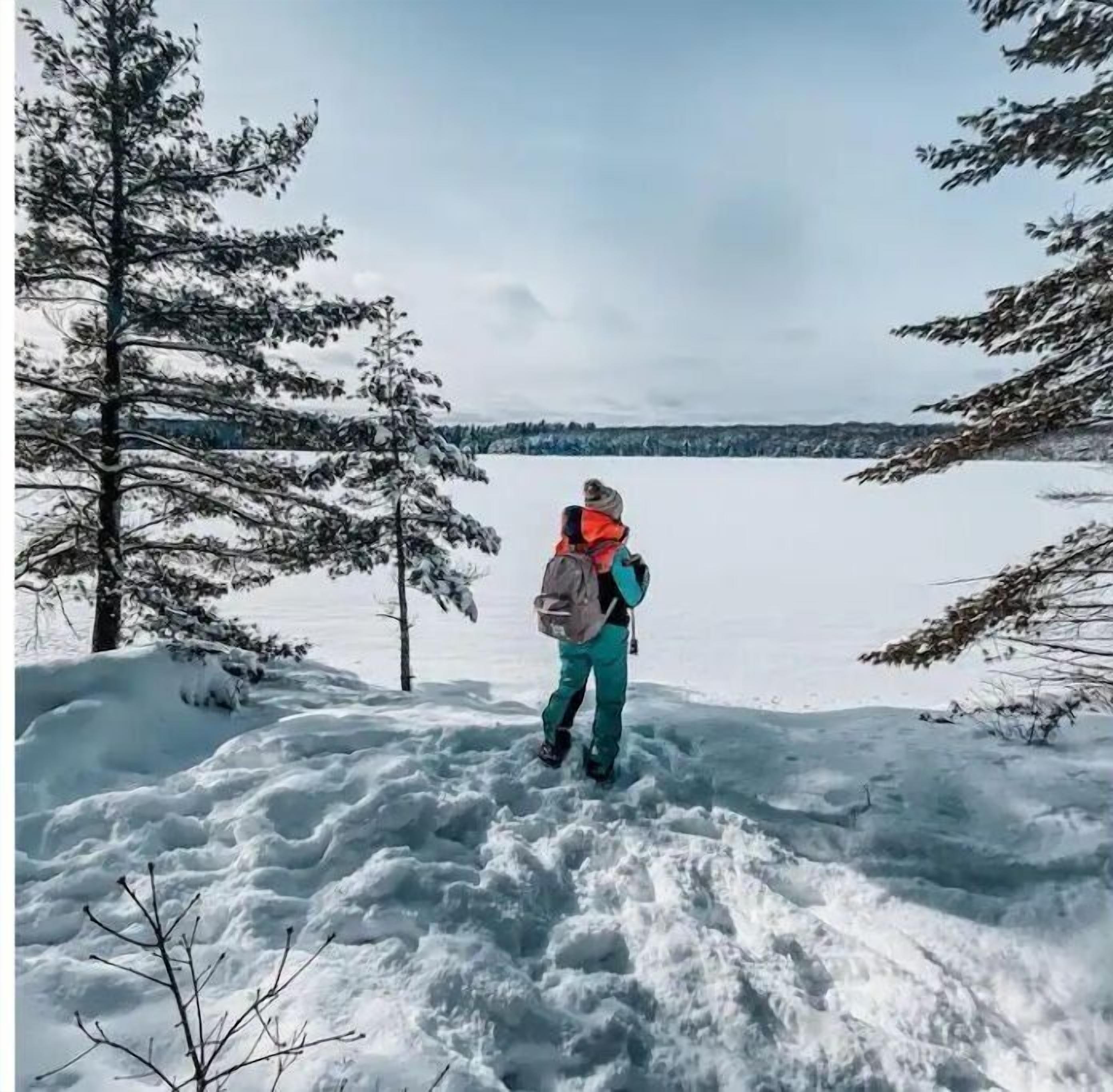 Une personne se tient sur un sentier enneigé, de part et d’autre se dressent des arbres chargés de neige, et elle contemple un paysage hivernal.