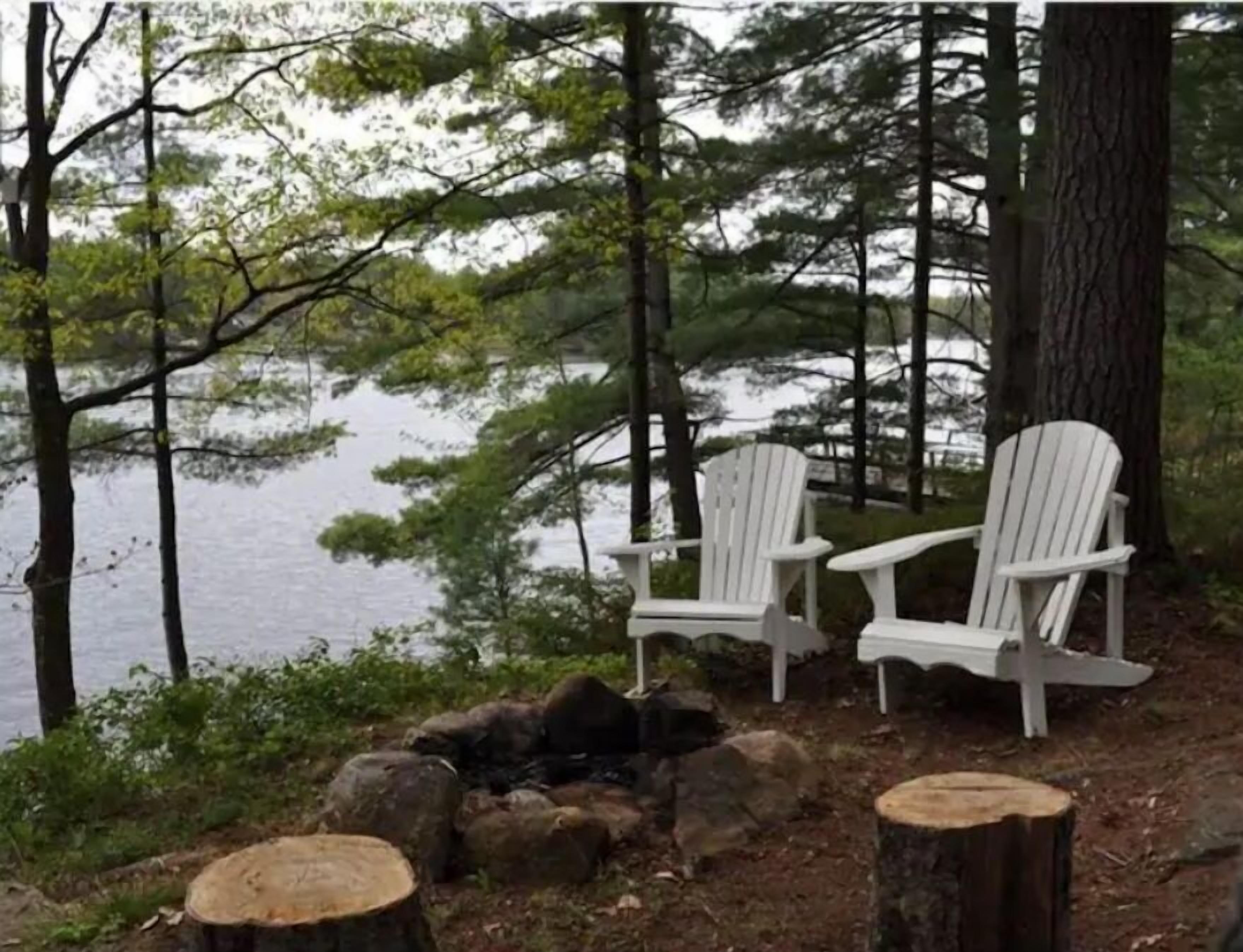 Two Adirondack chairs by a lake surrounded by trees.