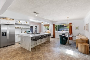 A modern kitchen with a central island, bar stools, a refrigerator, and a dining area with a table and chairs.