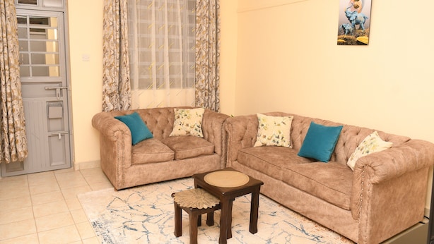 A living room with a brown sectional sofa, a small wooden coffee table, and a patterned rug. - Classic Beautiful 2-bedroom Apartment in Thika (Thika)