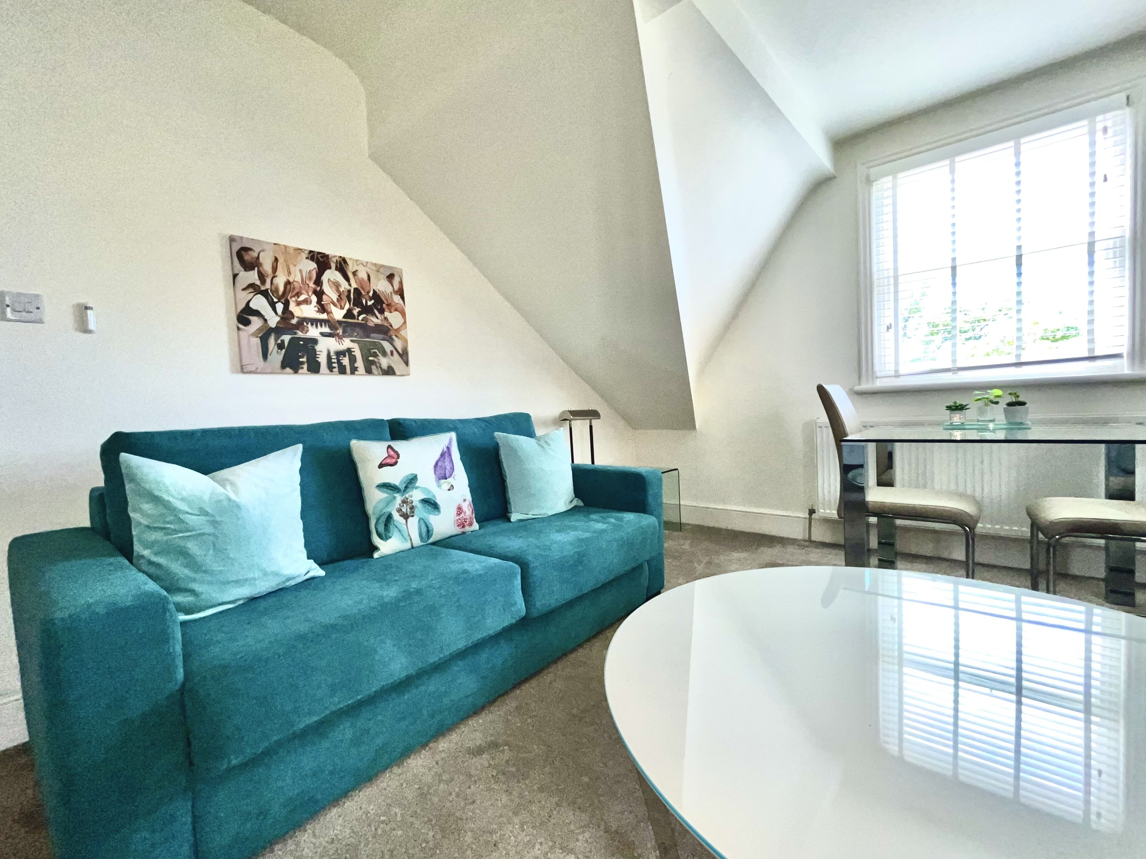 A modern living room with a teal sofa, a round glass table, and a large window with blinds.