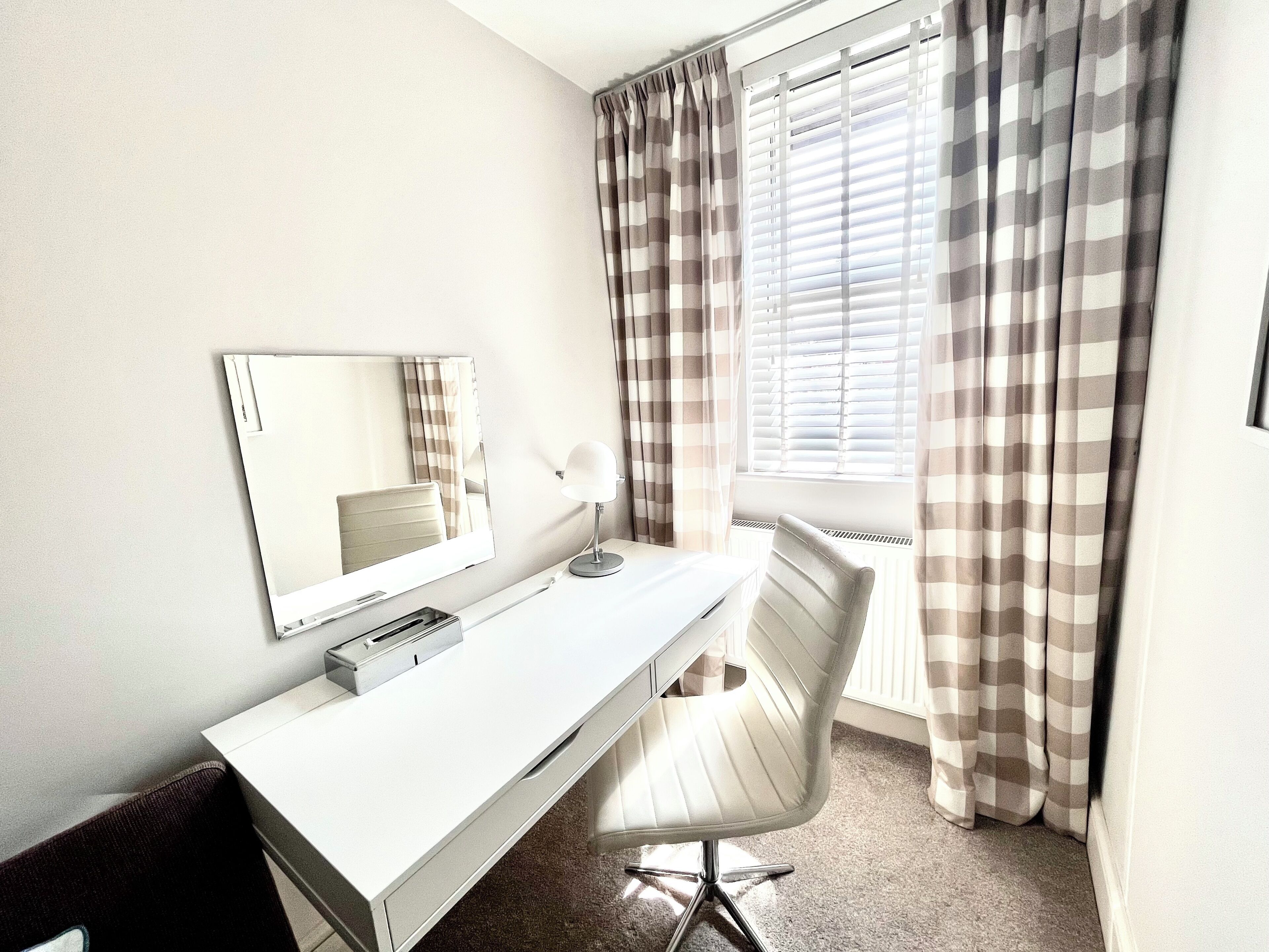 A hotel room with a white desk, a mirror, a lamp, a chair, and a window with blinds.