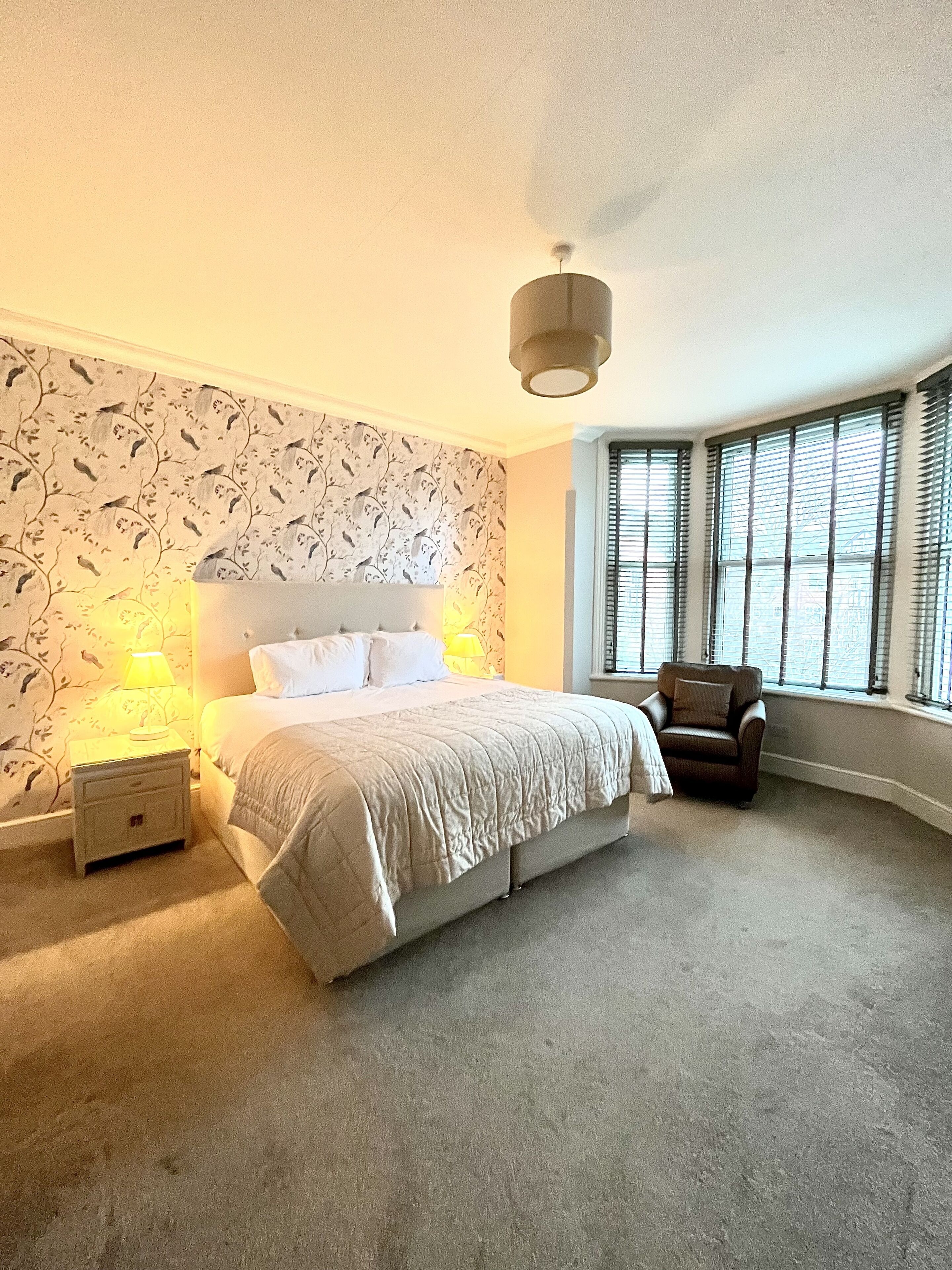 A bedroom with a large bed, a nightstand, a chair, and a window with blinds.