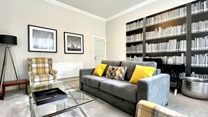 A living room with a grey sofa, yellow cushions, a plaid armchair, a glass coffee table, and a bookshelf filled with books. - Mode Apartments St Annes (Lytham St. Anne's)