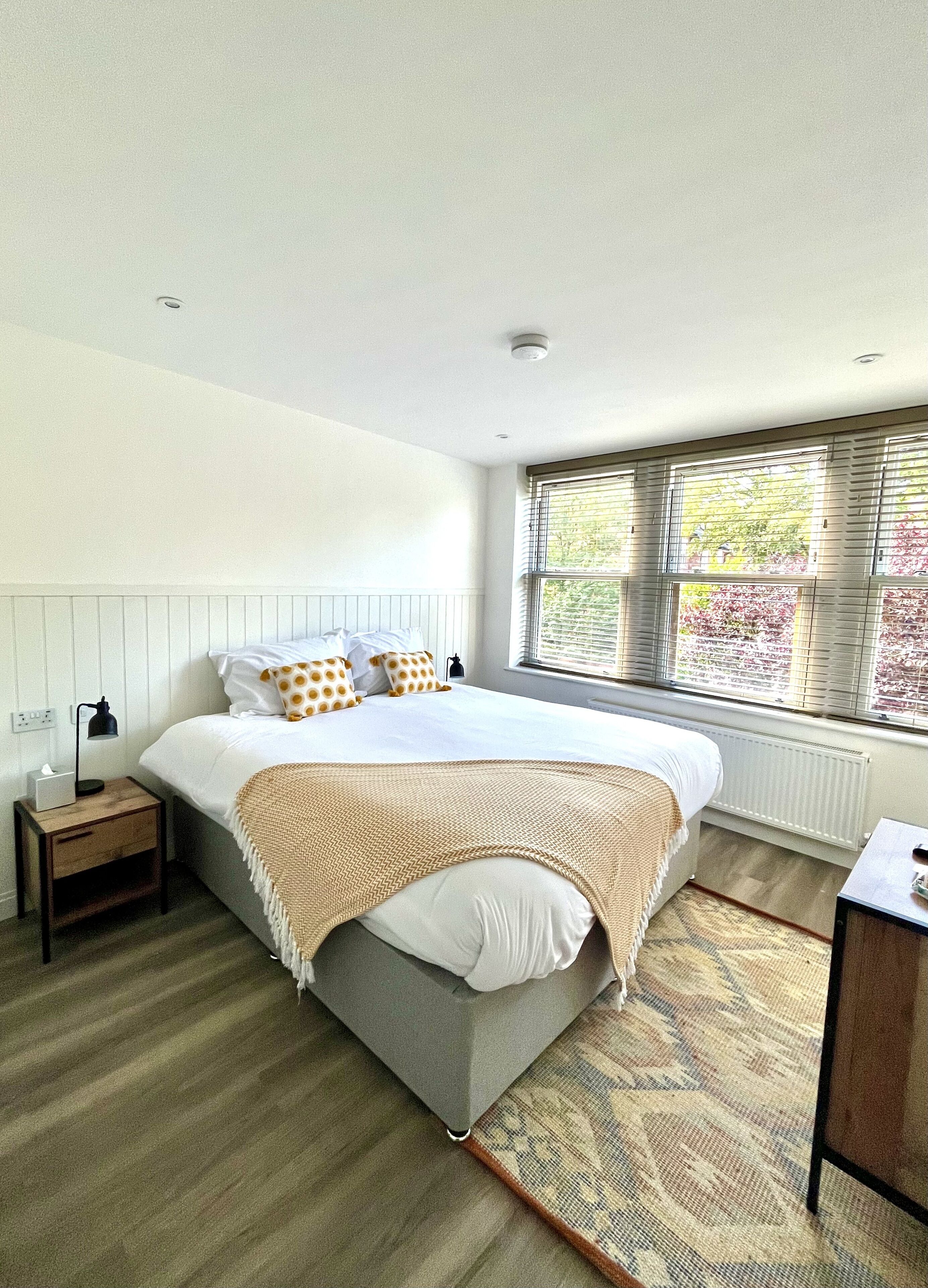 A bedroom with a bed, bedside table, lamp, window with blinds, and a radiator.