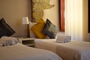 Quarto de hotel com duas camas, uma mesa de cabeceira, uma luminária e uma parede com textura.