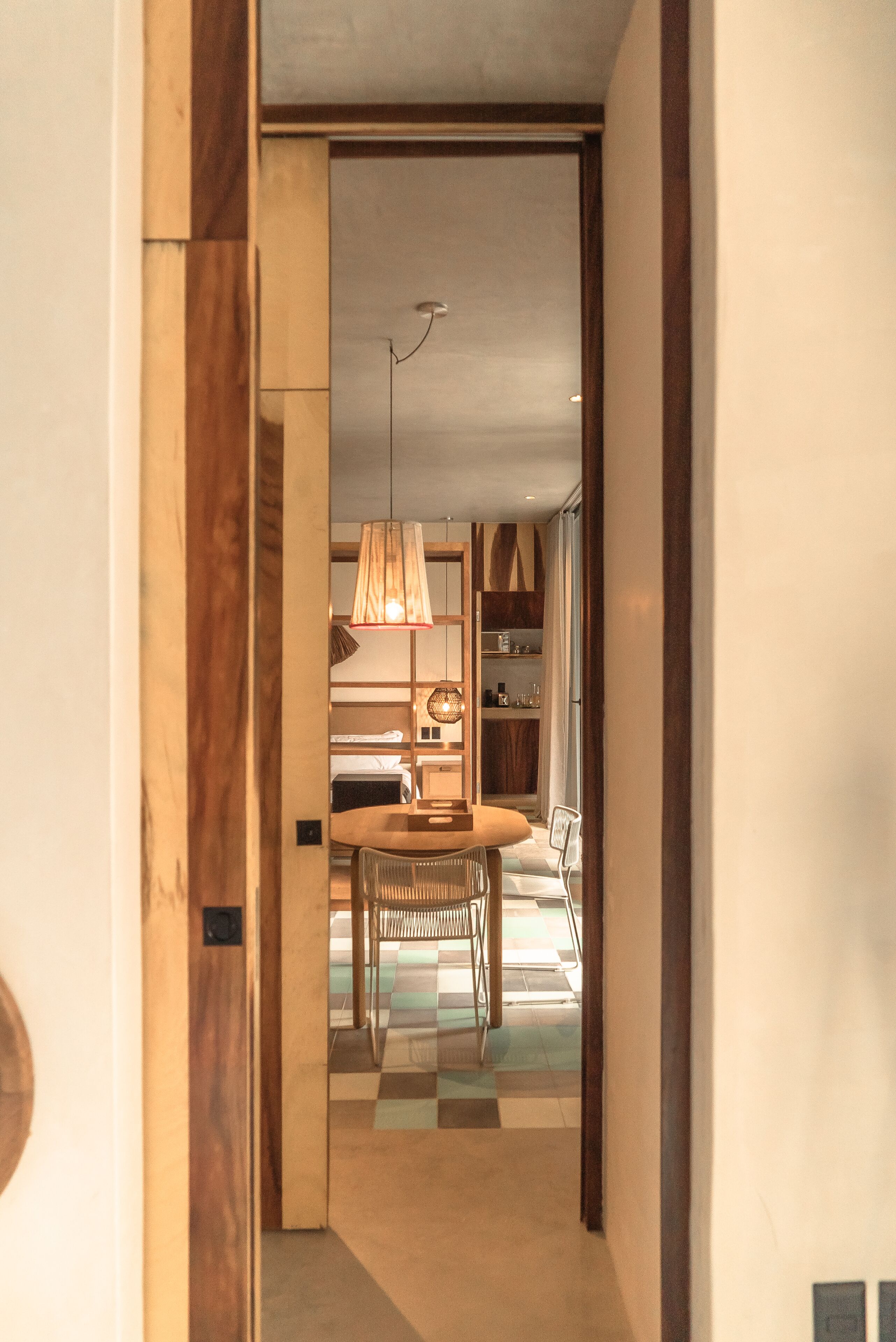 A hallway leading to a dining area with a checkered floor, a table, chairs, and a hanging lamp.