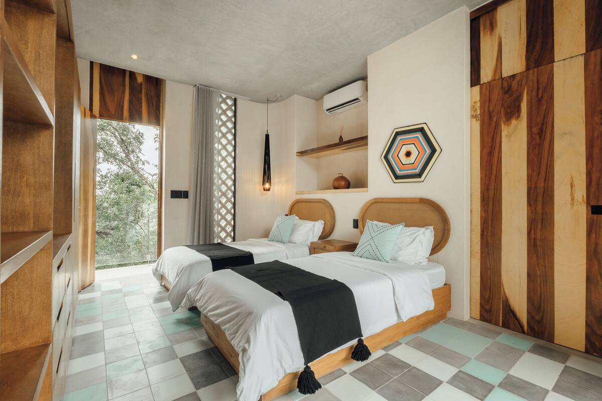 A bedroom with two beds, a window with a view, a geometric wall decoration, and a wooden wardrobe.