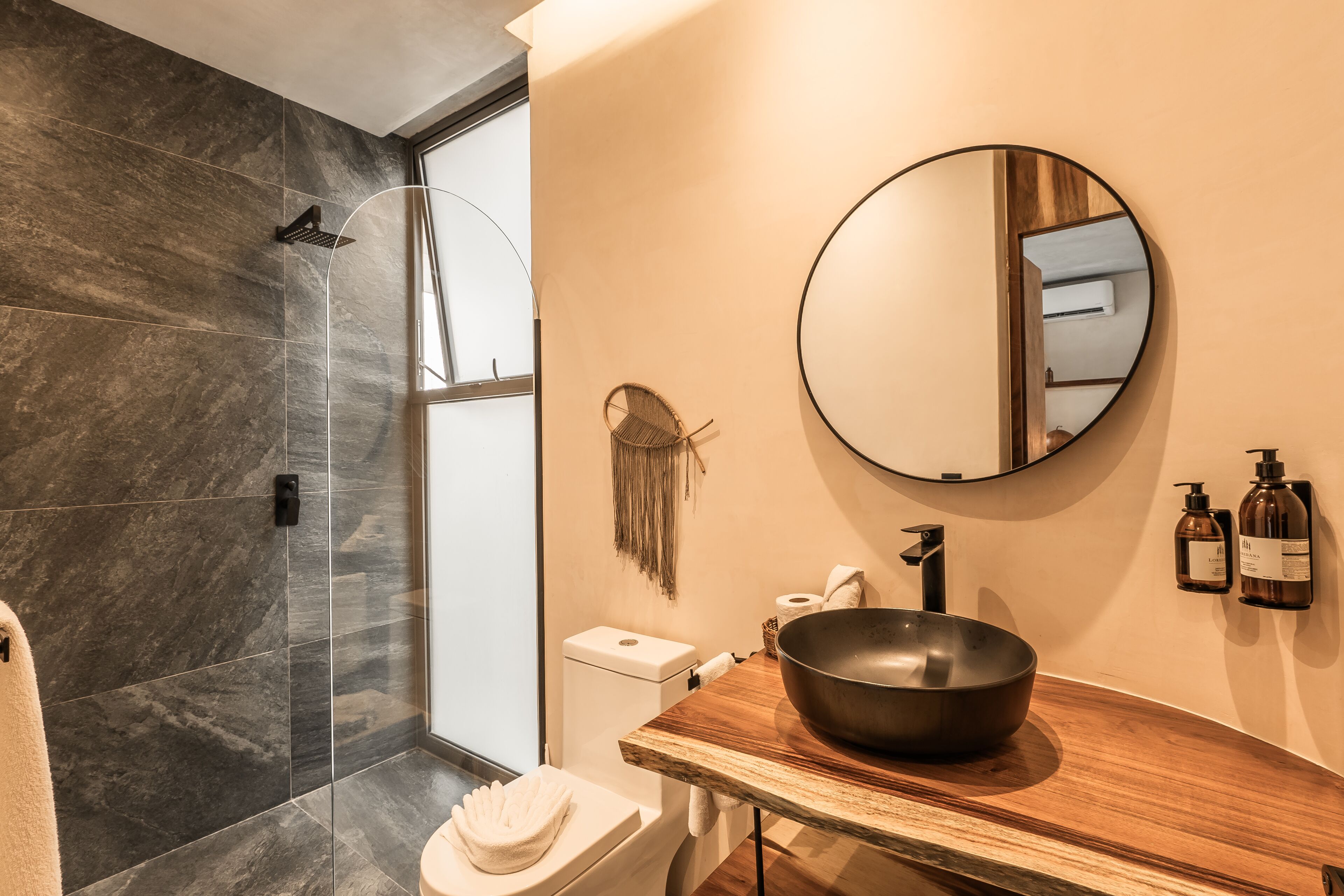 A modern bathroom with a large mirror, a wooden vanity, a black bowl sink, and a shower area with dark tiles.