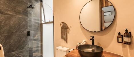 A modern bathroom with a large mirror, a wooden vanity, a black bowl sink, and a shower area with dark tiles.
